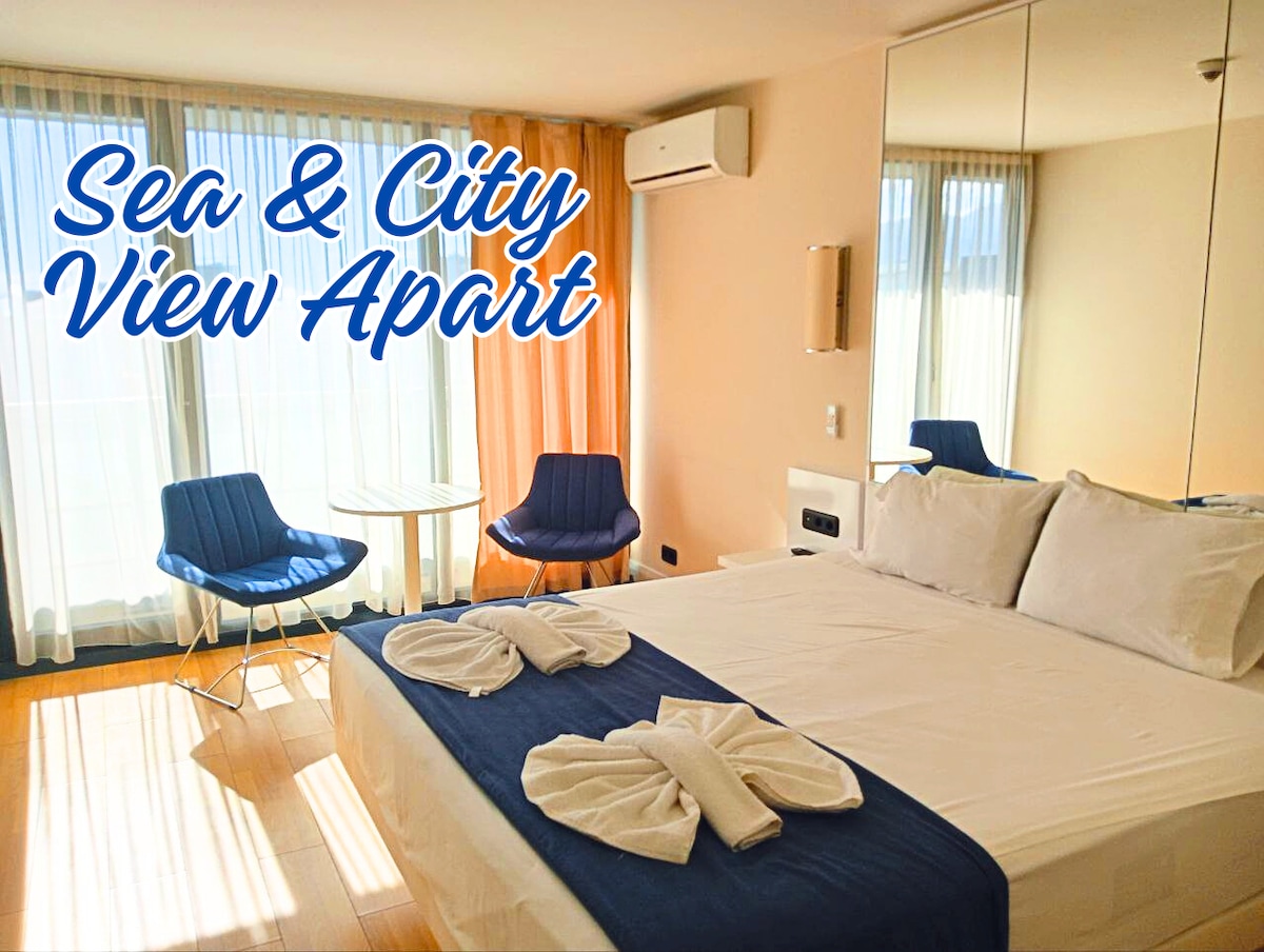 A cozy room features a spacious bed dressed in crisp linens, with decorative towels arranged on top. Two blue chairs and a glass table are positioned beside a large window that invites natural light, offering views of the sea and city beyond.