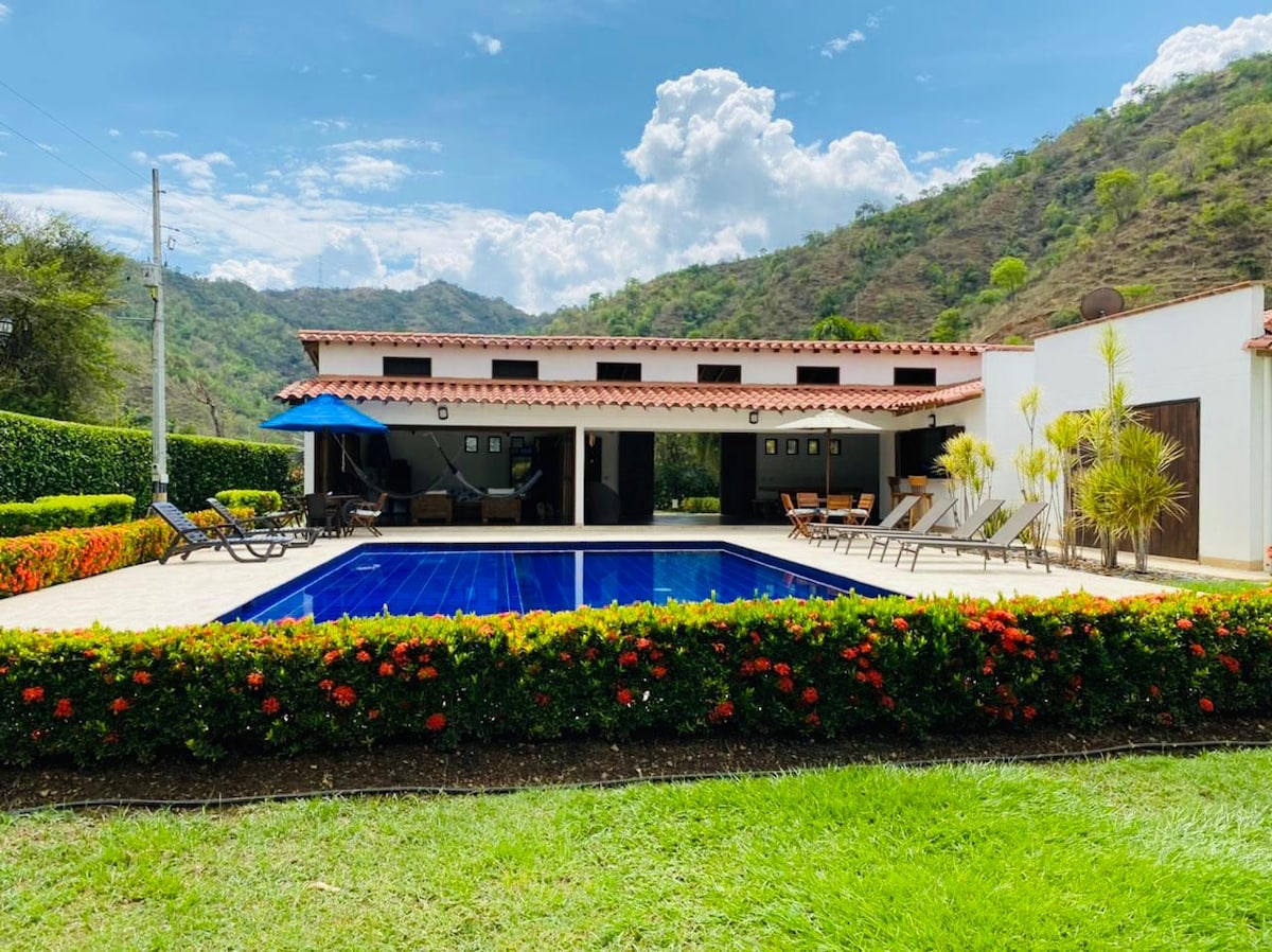 The exterior features a serene swimming pool surrounded by colorful flowerbeds and lounge chairs. A two-story building provides shaded areas with blue awnings, set against a backdrop of rolling green hills and a partly cloudy sky.