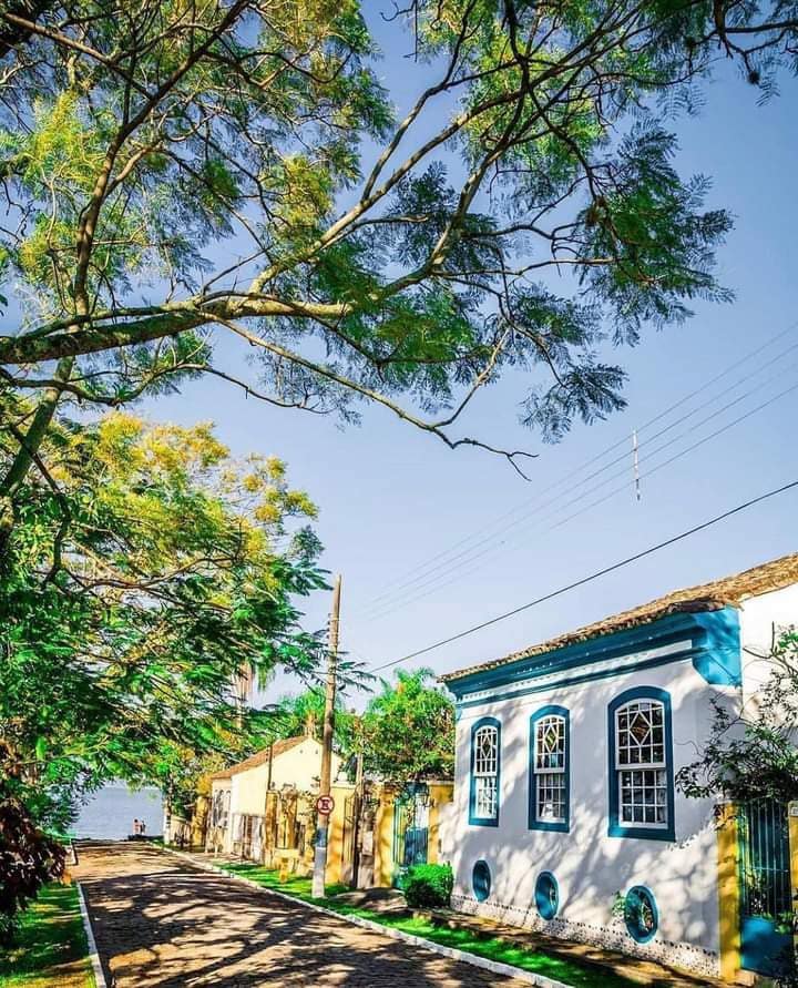 The image captures a quiet street lined with trees, providing shade to the charming buildings along the road. A blue and white structure is prominently featured, complemented by vibrant greenery. The distant view reveals a body of water, enhancing the scene's peaceful ambiance.
