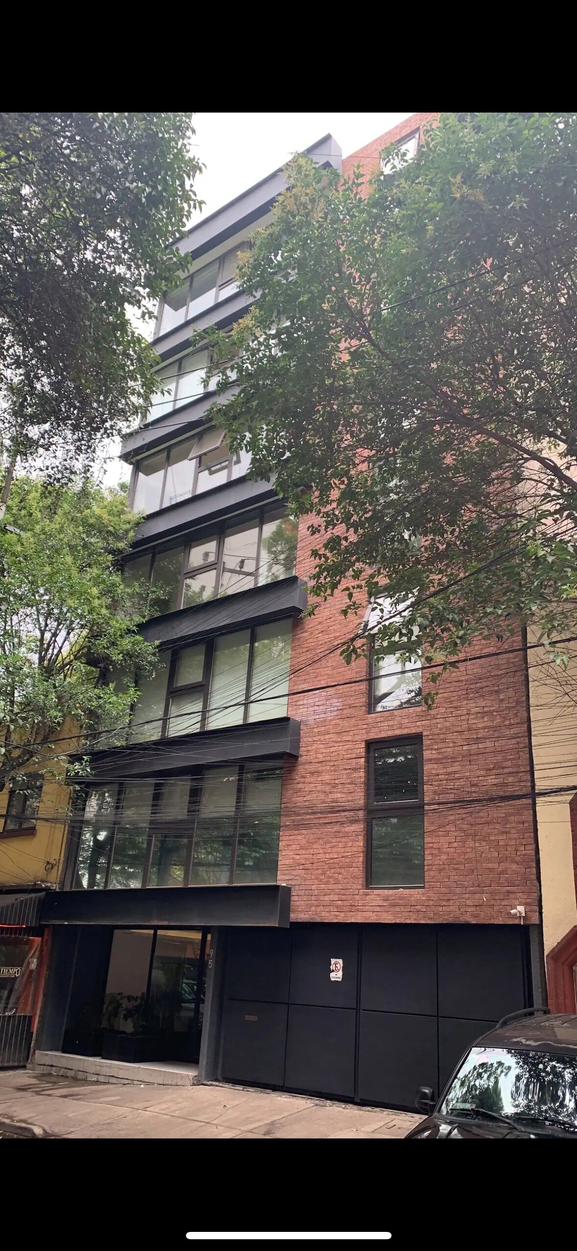 The building's exterior showcases a modern design with a combination of brick and glass elements. Large windows allow for natural light, while trees provide greenery, creating a balanced urban environment. The entrance is framed by a sleek, dark façade.