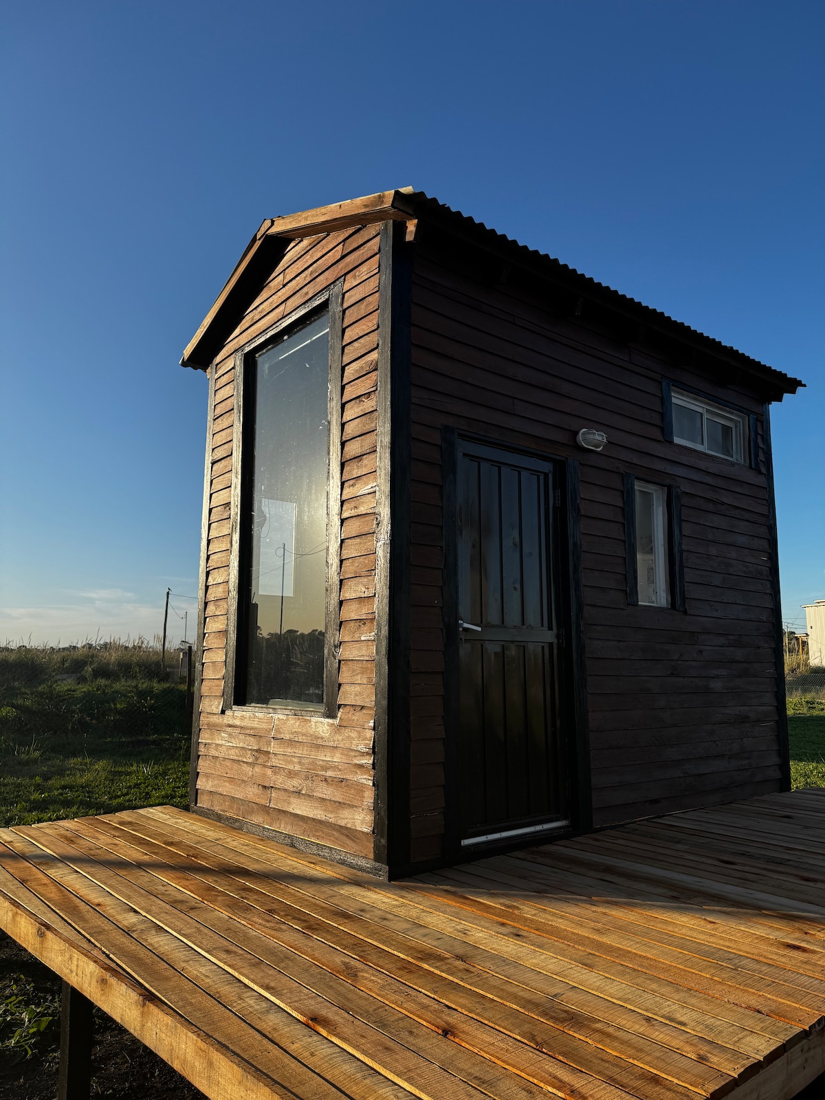 A tiny wooden house is presented against a clear blue sky. Large windows invite natural light, and a sturdy door offers access. The structure is set on a wooden deck, surrounded by open green fields, promoting a sense of privacy and tranquility.