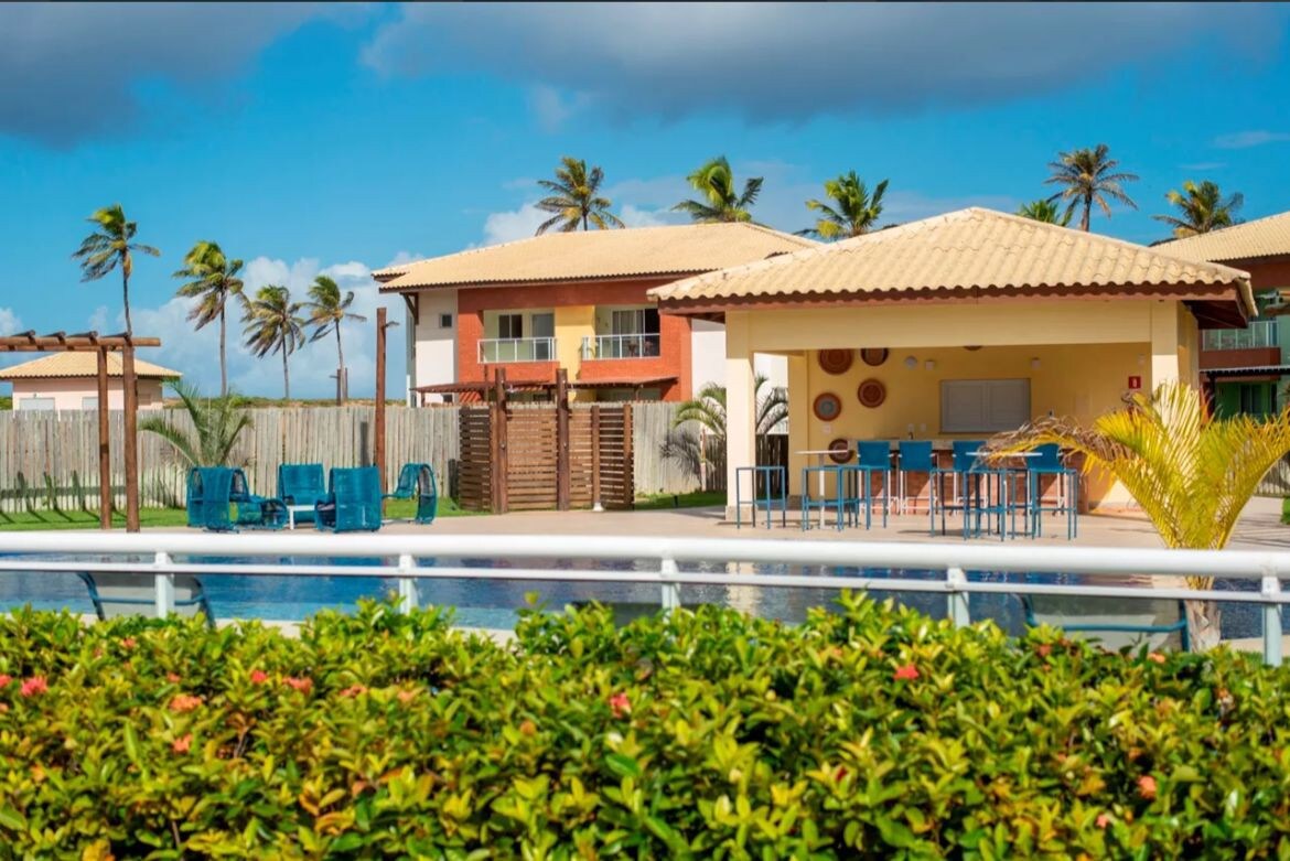A serene pool area is framed by lush greenery, with comfortable lounge chairs arranged around the water. Nearby, a covered outdoor seating area is visible, equipped with bar stools and a table. The vibrant colors of the surrounding buildings and palm trees enhance the peaceful setting.