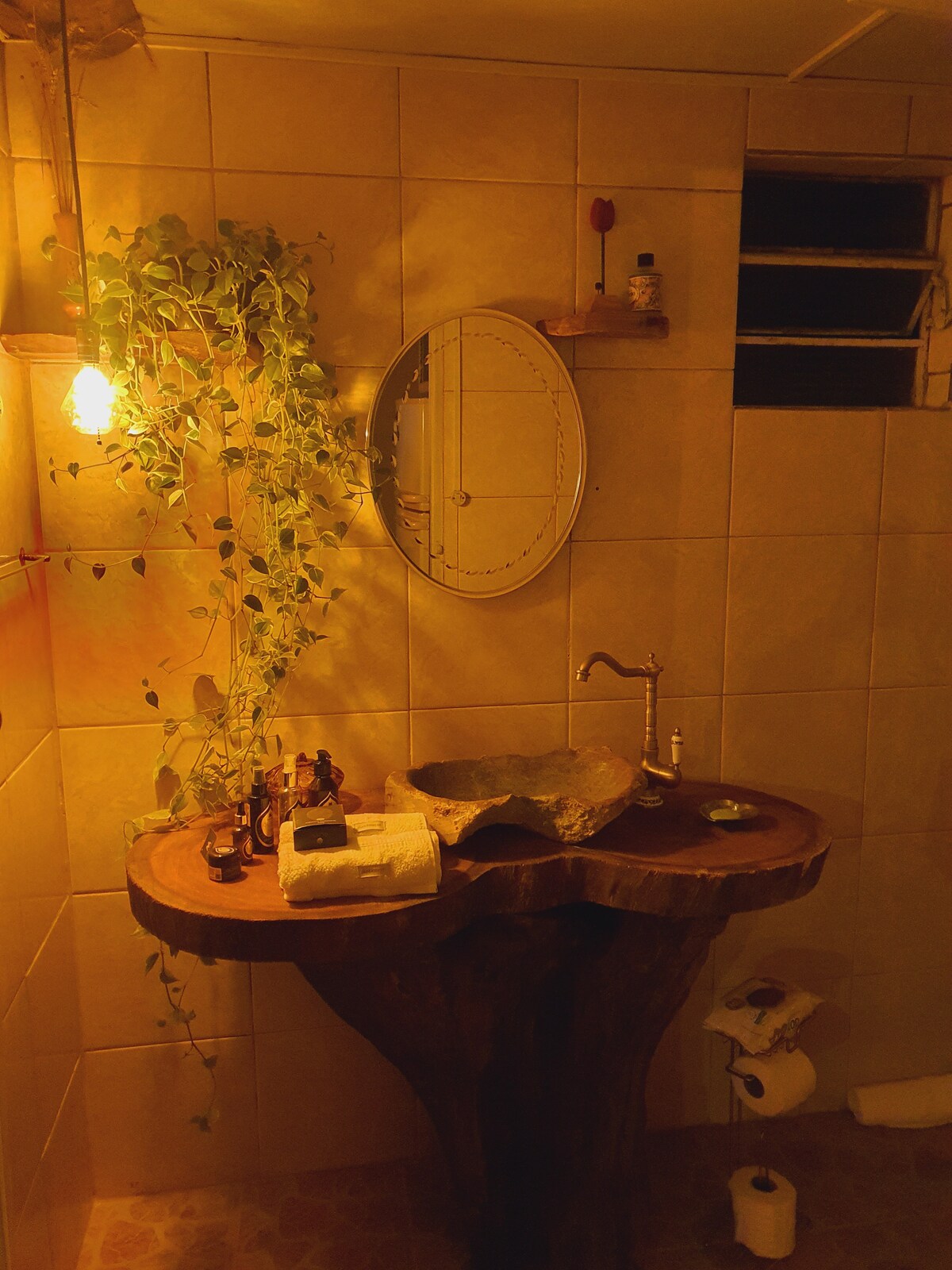 The bathroom features a unique wooden sink crafted from natural materials, positioned on a sturdy round table. A circular mirror is mounted above, while soft lighting creates a warm glow. A potted plant adds a touch of nature, enhancing the rustic ambiance.