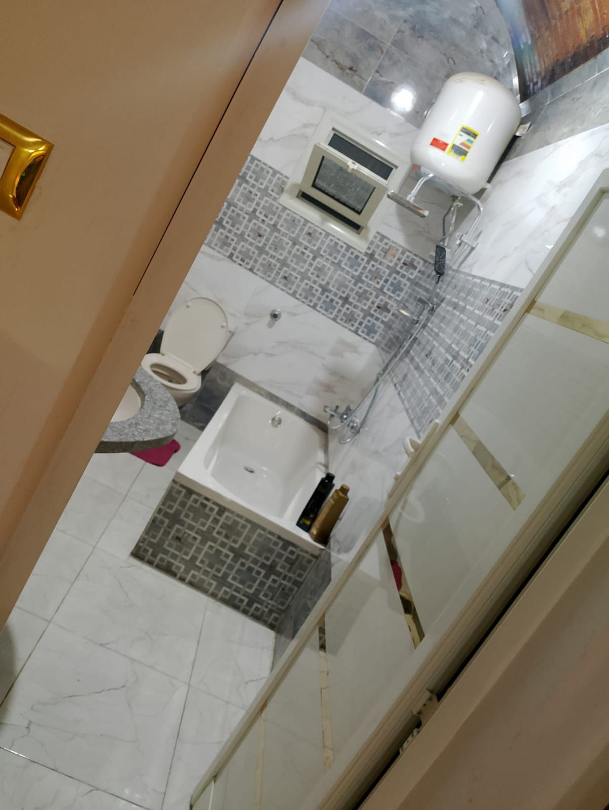 A modern bathroom is visible, featuring a bathtub integrated into a tiled platform. Grey and white tiles accentuate the walls. Basic fixtures include a toilet and a small window for ventilation, with a water heater mounted above the bathtub, ensuring practical amenities.