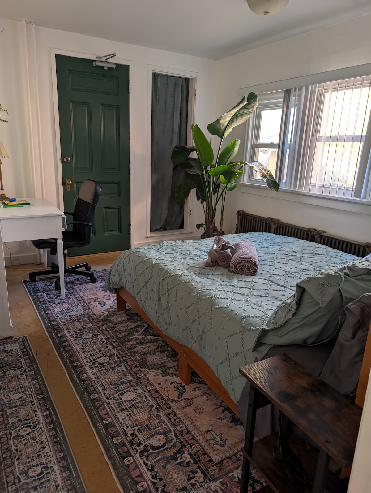 A serene bedroom features a spacious bed adorned with a textured green coverlet. A large plant adds natural decor beside the window, which is framed by two sets of blinds. A desk with a chair and a small nightstand completes the calming space.