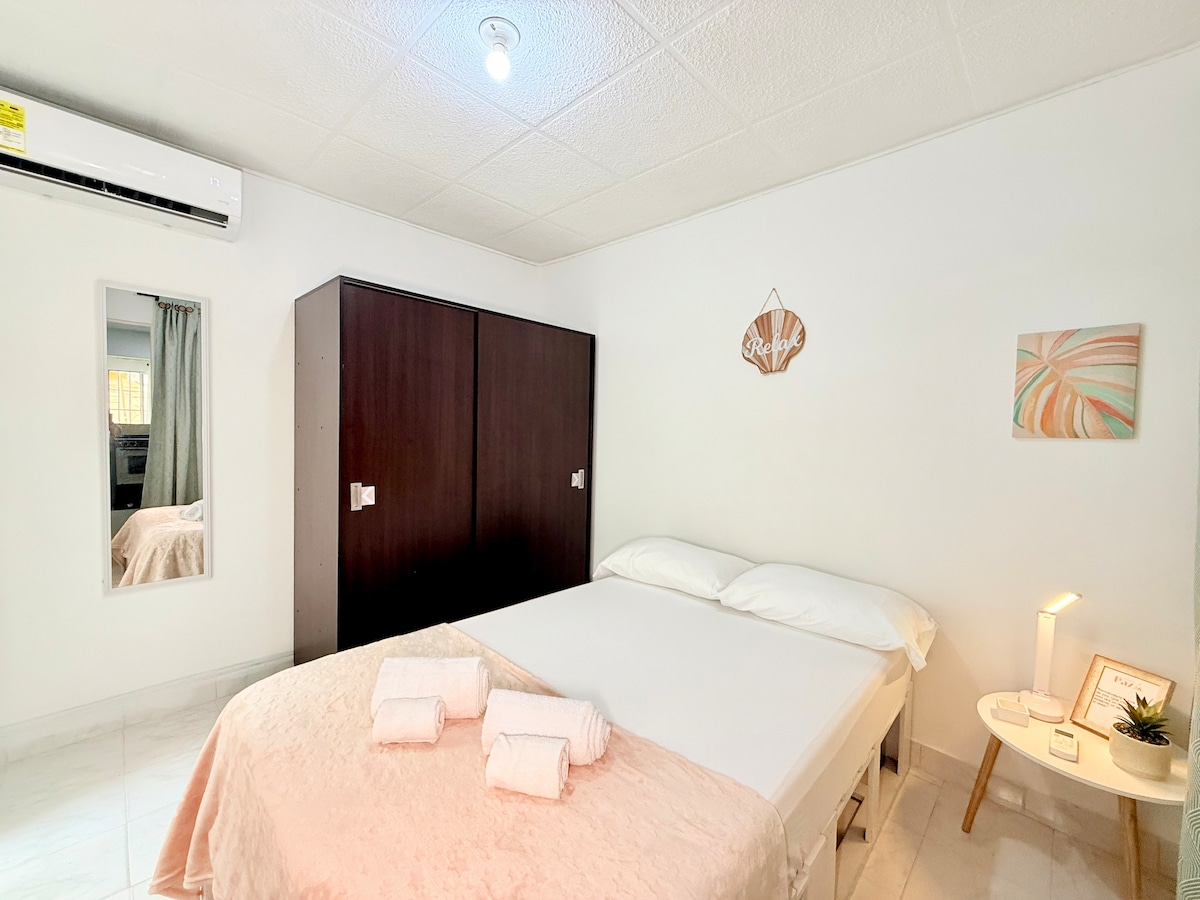A well-lit bedroom features a spacious bed with fresh white linens and plush towels displayed neatly. A mirrored wardrobe stands against one wall, while a modern art piece adds a splash of color above a bedside table that holds a lamp. Natural light filters in through the window.