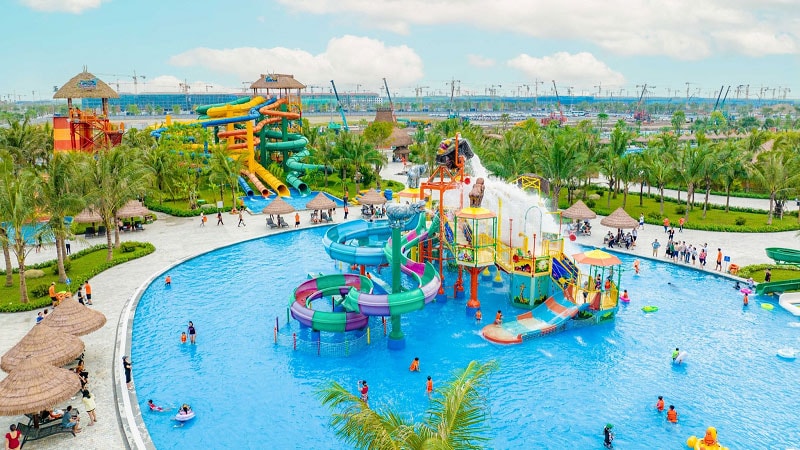 A vibrant water park is depicted, featuring multiple colorful slides and play structures surrounding a large, clear pool. Guests engage in various water activities. Lush palm trees are visible along the perimeter, and shaded cabanas provide relaxing spaces for visitors.