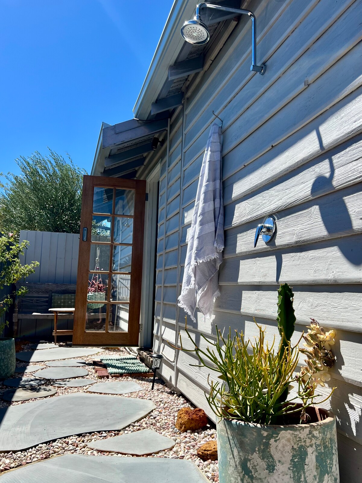 The image showcases a private outdoor space featuring a wooden door leading to a serene courtyard. A stone pathway is visible alongside plants in decorative pots. An outdoor shower with a modern fixture is mounted on the wall, enhancing the inviting ambiance.