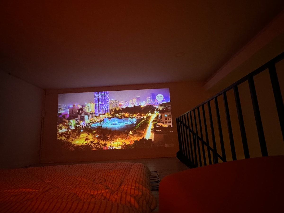 A living area features a spacious bed draped in a light coverlet, with a projector casting an image of Saigon's illuminated skyline on the wall. The scene highlights the city’s vibrant colors, while a sleek railing and a soft seating area add to the studio's contemporary design.