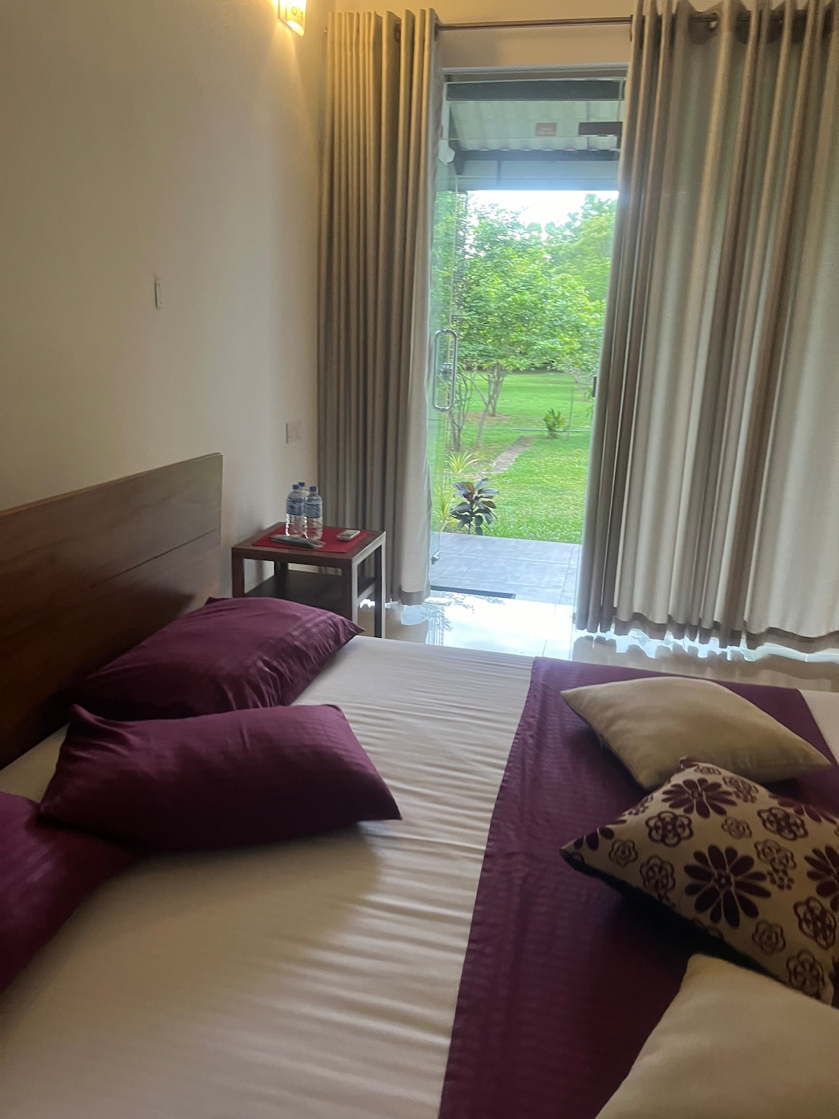 A comfortable sleeping area features a spacious bed adorned with purple and beige pillows. Natural light filters through large glass doors leading to a lush green outdoor space, complemented by light-colored curtains that frame the entrance.