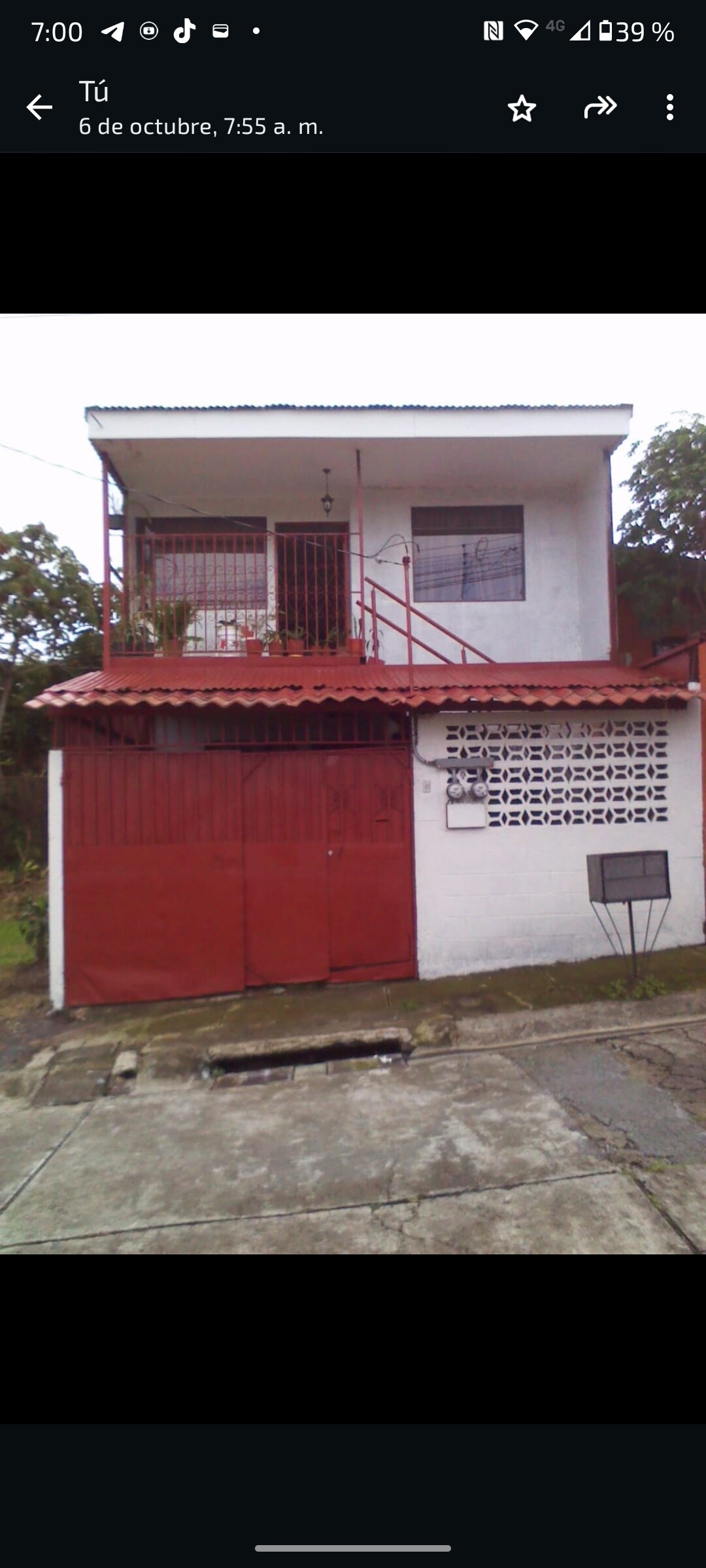 The building features two stories, with a red ground floor entrance and a white upper floor. The exterior has a brick accent wall and windows secured with grilles. A metal staircase leads to the second level, situated on a residential street with greenery surrounding the property.