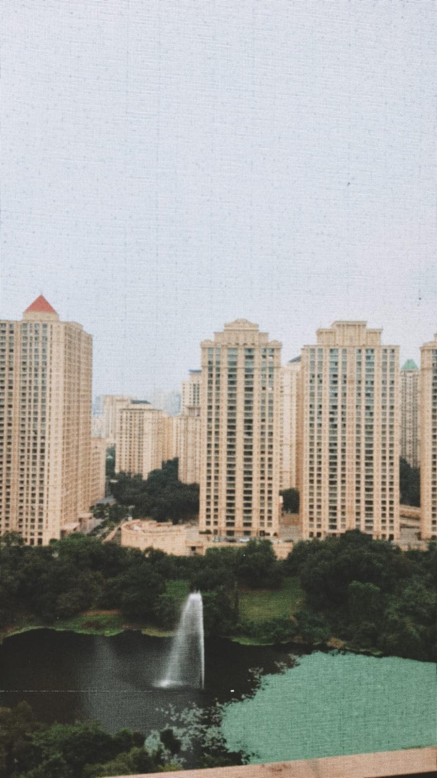A view of modern high-rise buildings surrounding a serene lake, featuring lush greenery and a fountain. The gentle water spray adds a calming element to the landscape, creating a peaceful urban environment.