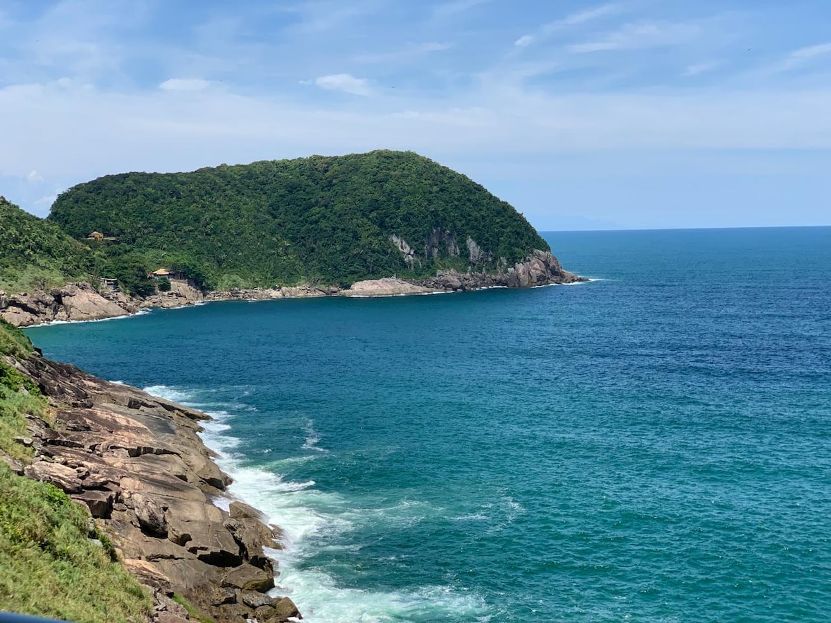 A panoramic view of the coastline reveals a lush, green hill rising from the blue ocean waters. Gentle waves are seen lapping against rocky shores, while the clear skies emphasize the vibrant colors of the landscape.