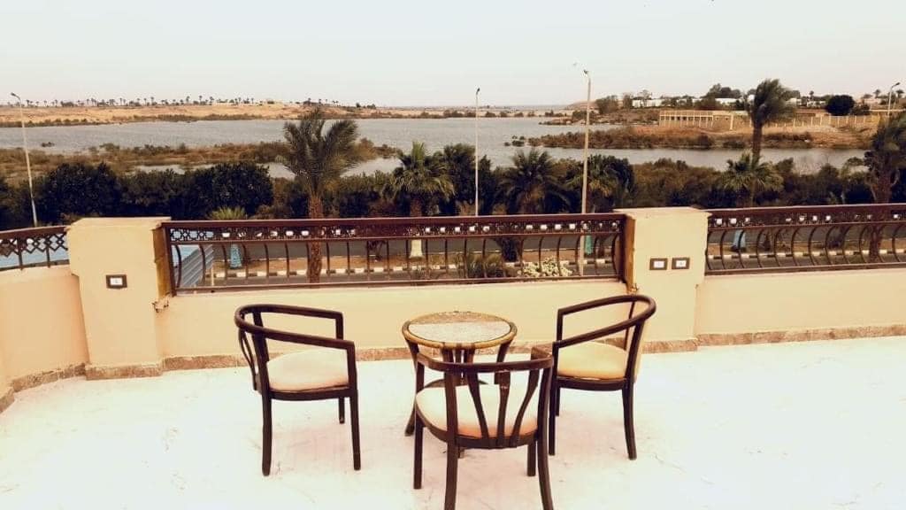 A terrace offers a view of the water, surrounded by greenery. Two chairs and a small table are positioned on the smooth, light-colored surface, inviting relaxation. The landscape includes trees and distant hills, contributing to a peaceful atmosphere.