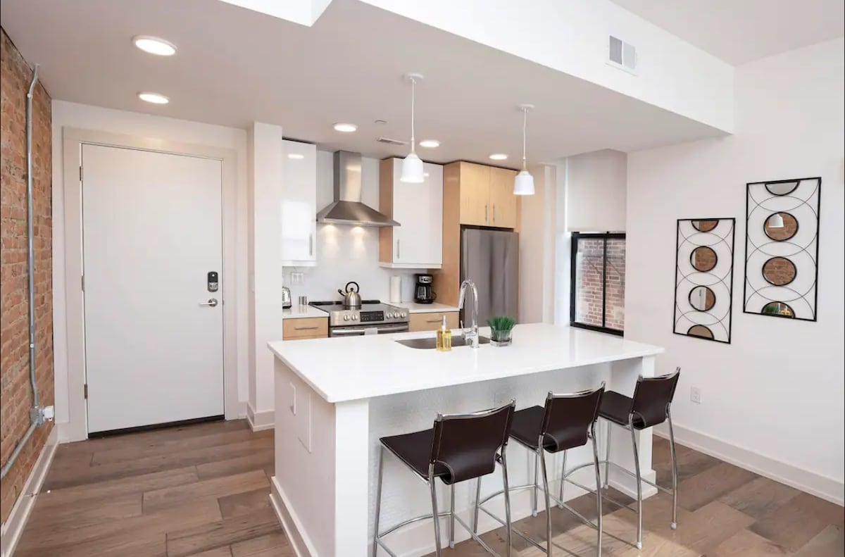 The kitchen features modern stainless steel appliances and quartz countertops, complemented by an island with three black chairs. Exposed brick adds character to the space, while pendant lights contribute to the overall brightness. A door leads to the entryway, enhancing accessibility.