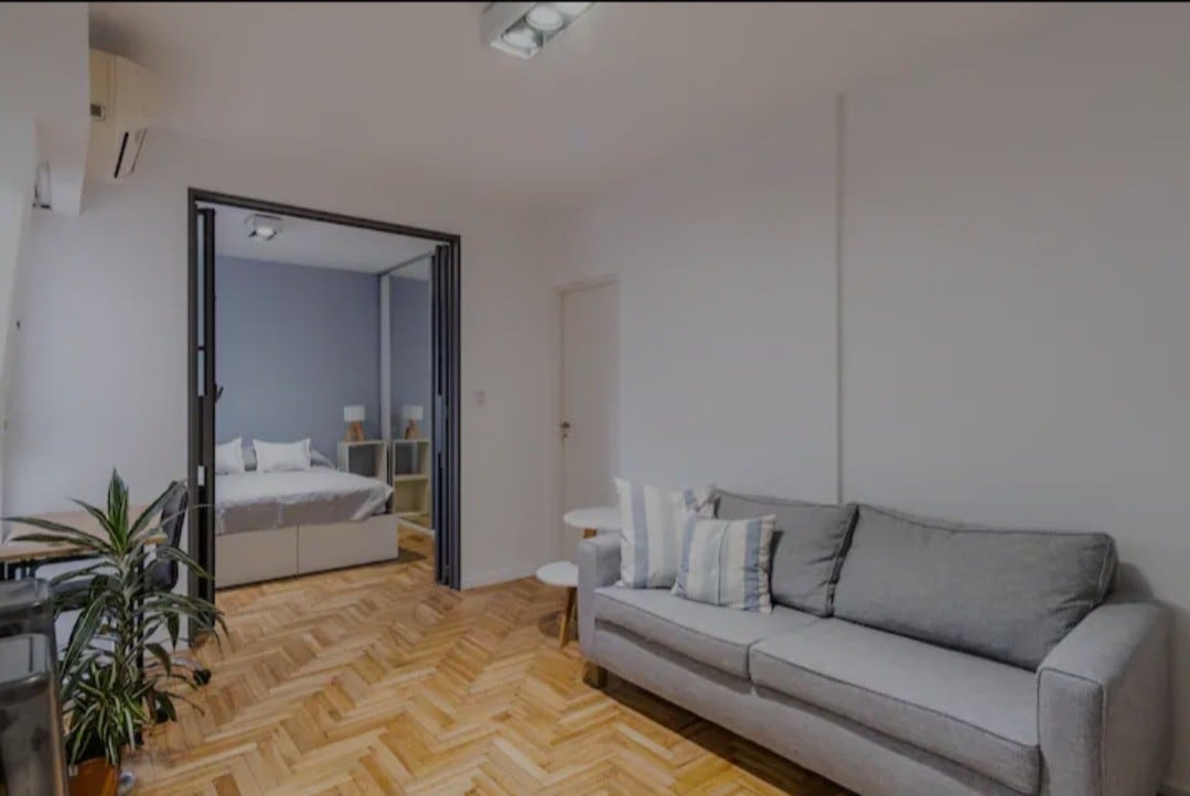 A living area features a comfortable gray sofa, complemented by a small side table. Natural wood flooring adds warmth to the space. An open doorway reveals a bedroom area with a spacious bed, enhancing the flow between rooms.