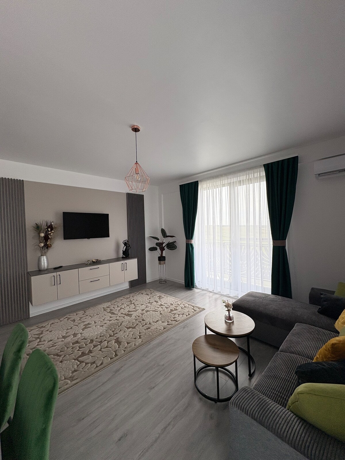 A modern living area is featured with a comfortable sofa and a pair of small tables. Natural light floods the space through sheer curtains, while a television is mounted on the wall. Decorative elements include a plant and vases, adding a touch of elegance.