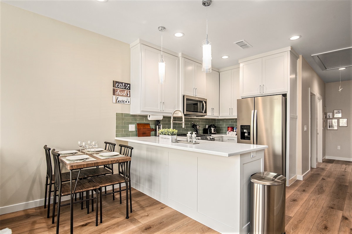 A modern kitchen is featured with sleek white cabinetry and stainless steel appliances. A dining table with four black chairs sits adjacent, set with tableware. Bright pendant lights illuminate the space, while a small herb garden is visible on the countertop, adding a natural touch.