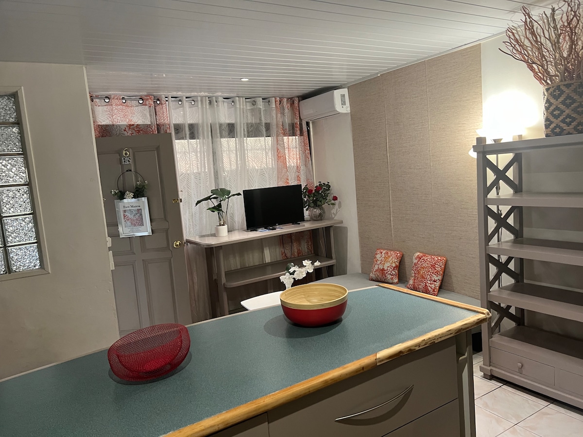 The welcoming living area features a light-colored wall with decorative accents and cozy seating. A wooden shelf holds ornamental pieces, while a television is positioned on a console. Natural light filters through sheer curtains, creating a bright environment for relaxation.
