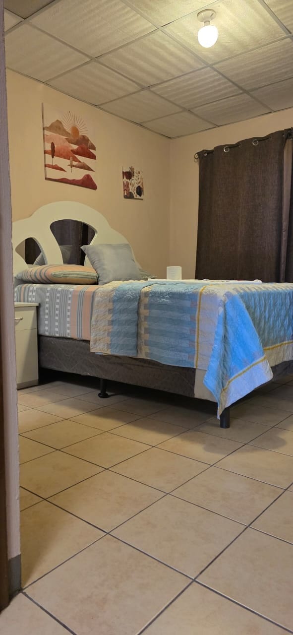 A cozy bedroom features a spacious bed with a patterned blue and white blanket. Soft light filters in from a nearby window, partially covered by dark curtains. Wall art adds a touch of color to the neutral walls, and tile flooring provides a clean, modern look.