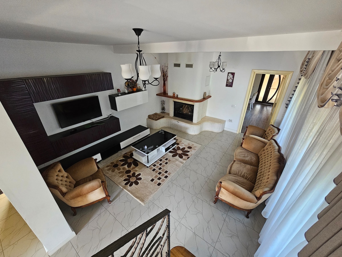 A spacious living area features a cozy seating arrangement on plush sofas, arranged around a central coffee table. A subtle area rug with floral designs lies beneath. A modern entertainment unit is mounted on the wall, and a fireplace adds warmth to the room.