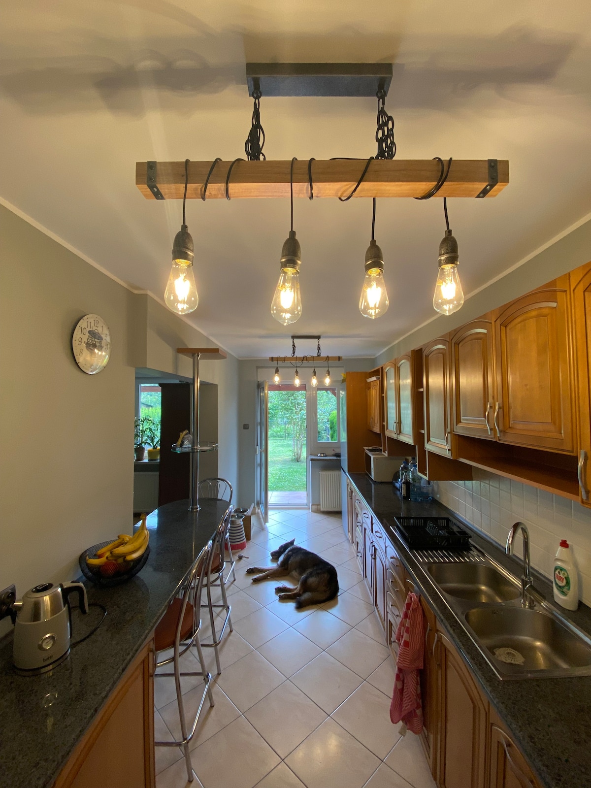 A spacious kitchen features wooden cabinetry and a long, dark countertop. Multiple bar stools line the counter, complementing the hanging light fixture with exposed bulbs. In the background, a doorway leads to a garden area, while a dog rests on the floor nearby.