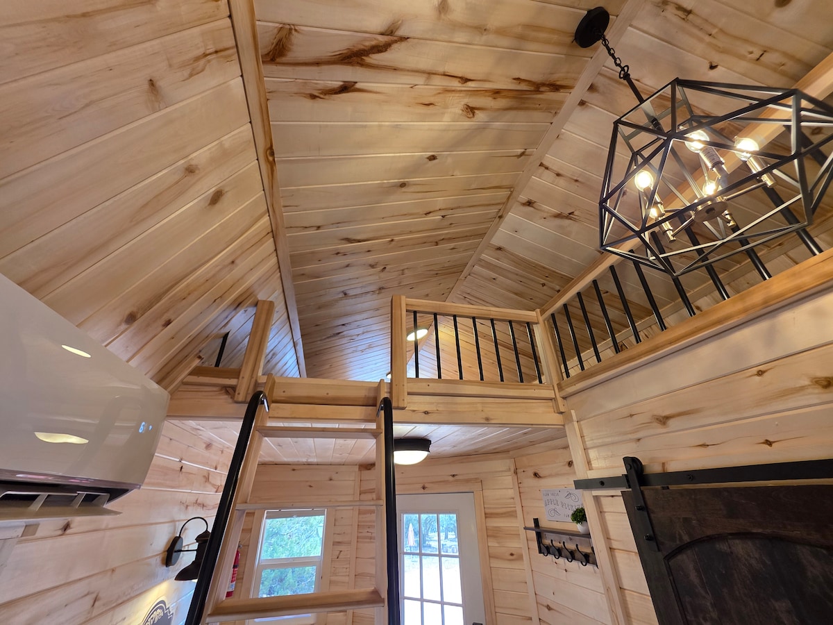 The interior of the cabin features warm wood paneling and a high ceiling. A staircase leads to a loft area with railings, and a metal light fixture hangs from above. Large windows provide natural light, enhancing the cozy atmosphere.