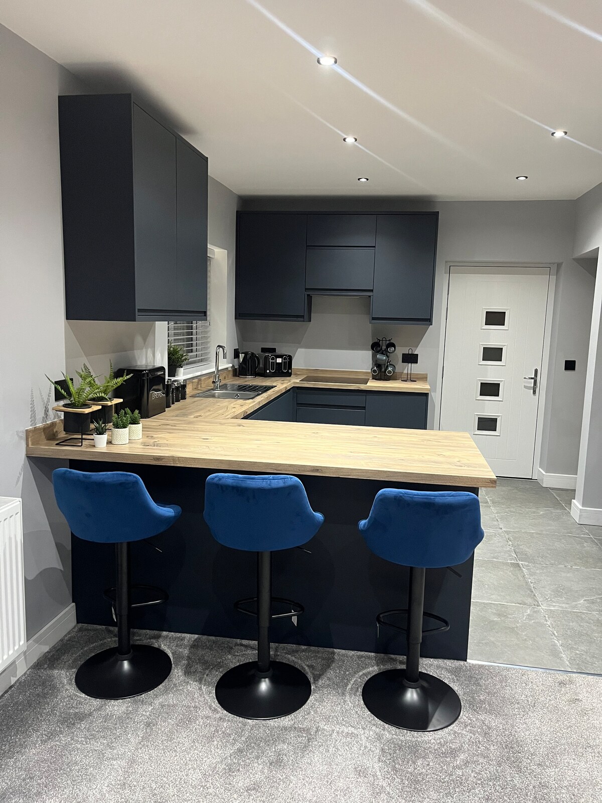A modern kitchen is presented with sleek dark cabinetry and a light wooden countertop. Three blue upholstered stools are positioned at the island, offering a casual dining space. Natural light floods the room, accentuating the contemporary design and grey tiled flooring.