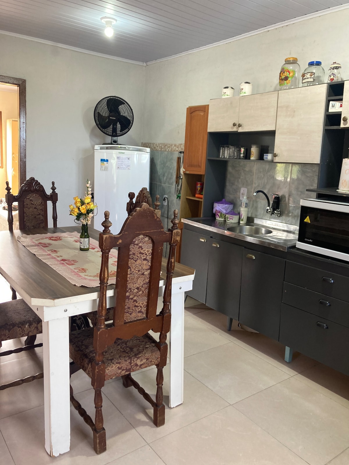 A well-equipped kitchen features a wooden dining table surrounded by high-backed chairs. A refrigerator and microwave are visible, along with countertop appliances. The cabinets showcase dishware and decorative items. Bright lighting highlights the functional layout and cheerful plants on the table.