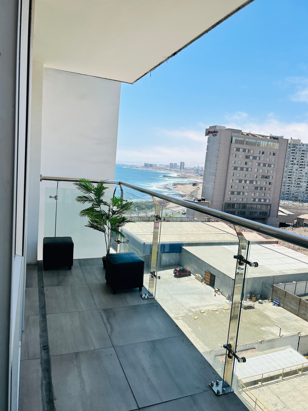 A balcony is featured with glass railings, allowing for unobstructed views of the coastline and nearby buildings. Two black seating cubes are placed on the tiled surface, complemented by a potted plant. Natural light fills the area, creating a pleasant outdoor space.