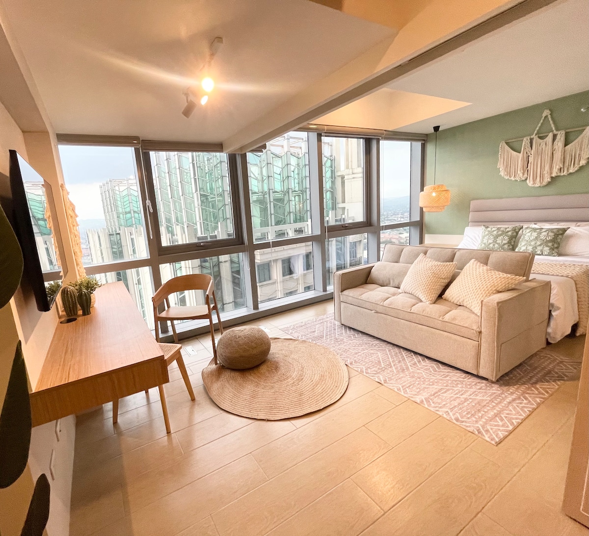 A cozy living area features a light-colored sofa and a wooden desk against a backdrop of large windows showcasing a city skyline. A round area rug and decorative pillows enhance the inviting atmosphere. The calming green walls add to the relaxing environment.