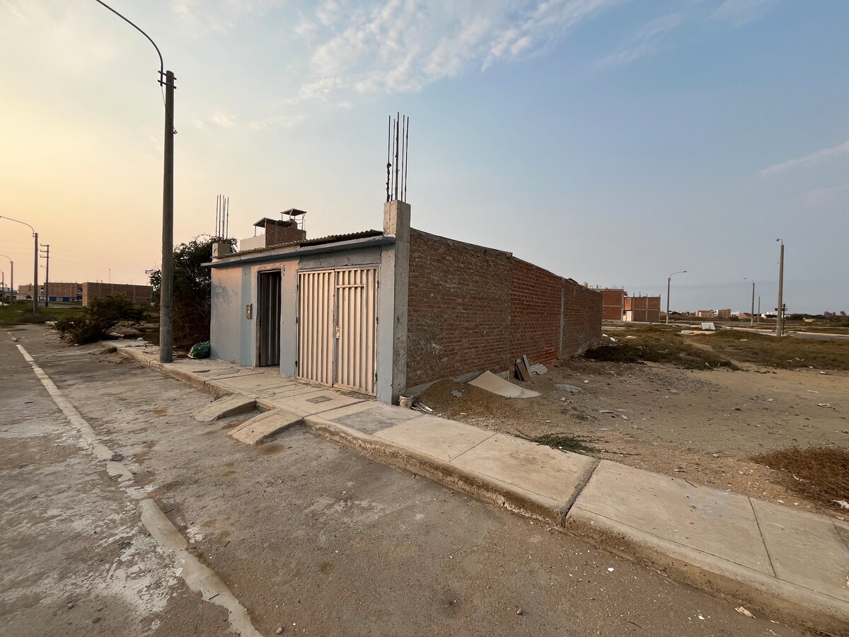 An exterior view of a simple, single-story building is presented, featuring a partially open door and a brick wall. Surrounding areas are mostly empty, with a concrete pathway leading up to the entrance. Utility poles are visible in the background under a soft evening sky.