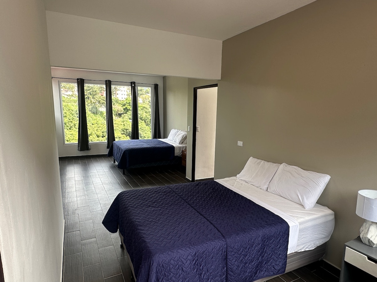 The room accommodates two beds with dark blue bedding, positioned to allow ample natural light through large windows. A minimalist design is featured on the walls, contributing to a spacious feel. A nightstand with a lamp sits beside one of the beds.