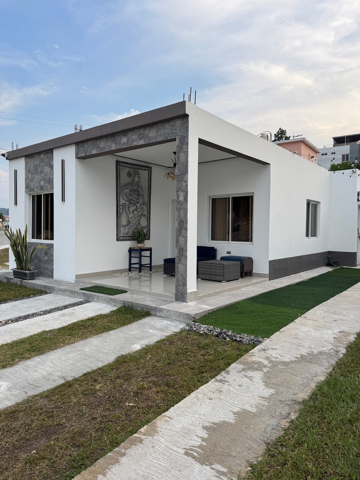 A modern single-story house features a clean white exterior with stone accents. The entrance is flanked by neatly manicured grass and a stone pathway. Large windows allow natural light into the living space, with a blue chair visible on the patio.