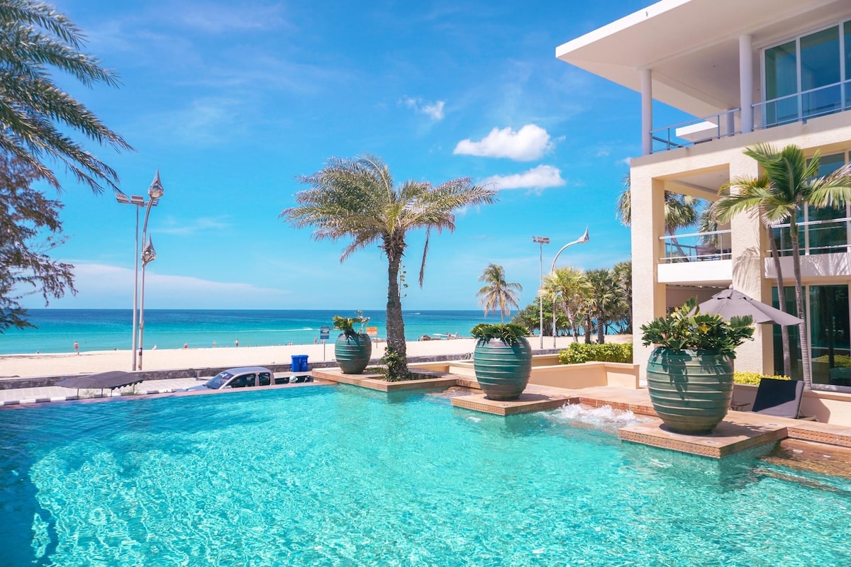 A tranquil pool area features shimmering turquoise water, surrounded by lush palm trees and decorative planters. The beach is visible in the distance, with soft waves meeting the sandy shore. A modern building is positioned nearby, enhancing the coastal ambiance.