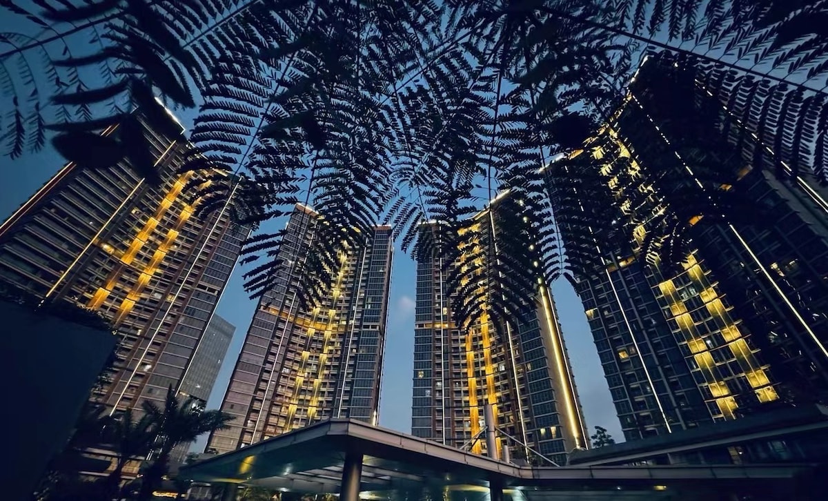 The modern high-rise buildings are framed by lush ferns, showcasing illuminated balconies and windows against the twilight sky. The sleek architecture reflects a contemporary style, highlighting the combination of greenery and urban design.