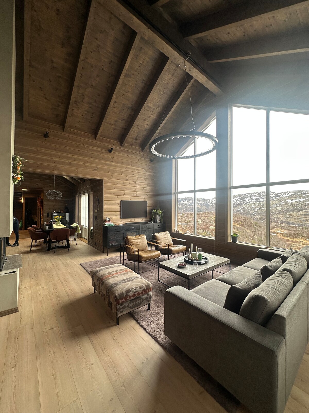 A spacious living area features high wooden ceilings and large windows offering scenic views. A comfortable sofa and patterned ottomans are arranged around a coffee table, while a TV is mounted on the wall. Natural light brightens the space, complementing the earthy tones of the decor.