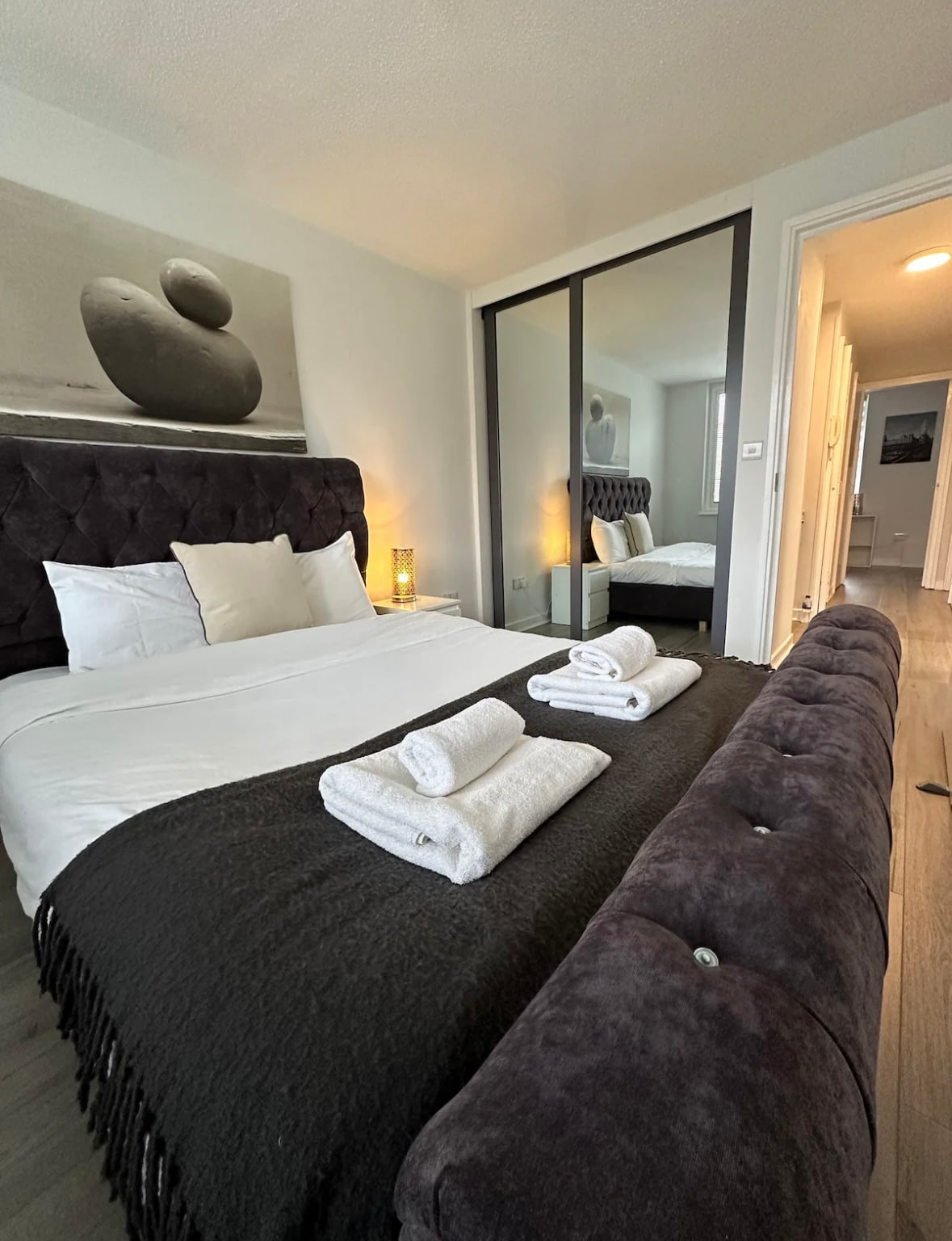 A well-appointed bedroom features a plush headboard with light-colored bedding neatly arranged. Soft towels are displayed at the foot of the spacious bed. A mirrored wardrobe reflects the room's layout, while soft lighting adds a touch of warmth to the modern decor.