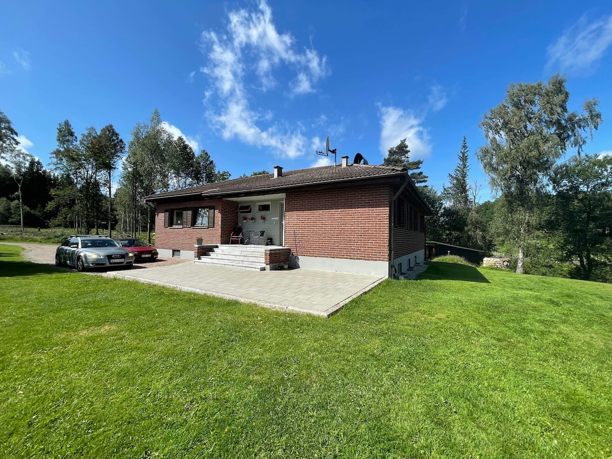 A modern brick house is set on a grassy lawn surrounded by trees. The exterior features a welcoming entrance with steps leading up to it, and a paved area accommodates a parked car. Clear skies add to the serene setting.