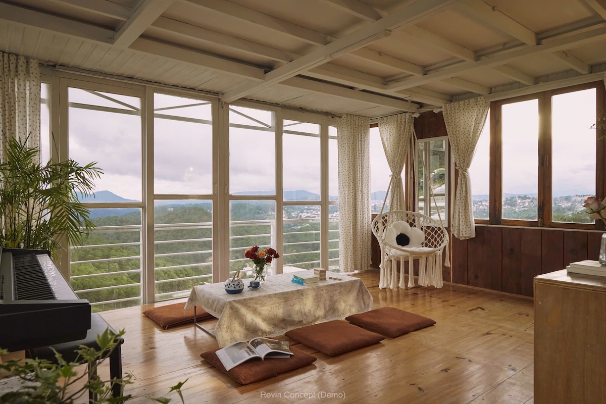 A bright living area features large windows that invite natural light, offering views of the surrounding landscape. Floor cushions are arranged around a low table, complemented by a cozy chair. Lush plants add a touch of greenery, enhancing the calm atmosphere.