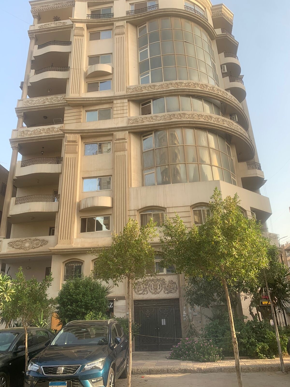 A modern building is showcased with a curved facade and large glass windows, allowing natural light to flood the interior. Lush greenery is present in the foreground, enhancing the inviting exterior. Vehicles are parked nearby, signifying easy accessibility.