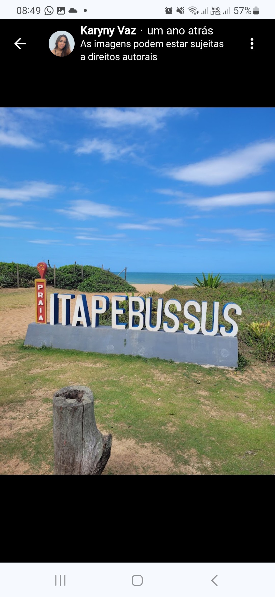 A large coastal sign reading 'ITAPE BUSSUS' is positioned on a sandy pathway leading towards the beach. Bright blue sky and gentle waves are visible in the background, complemented by greenery and shrubs surrounding the area.
