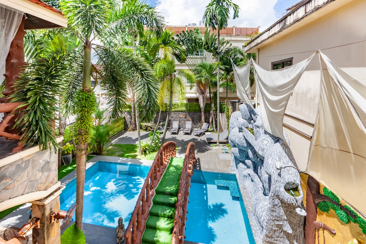 A serene outdoor space is visible, featuring a turquoise pool bordered by lush greenery. Comfortable lounge chairs are arranged on a patio area, while a decorative stone feature adds charm. A partial view of a shaded area provides an inviting spot for relaxation.