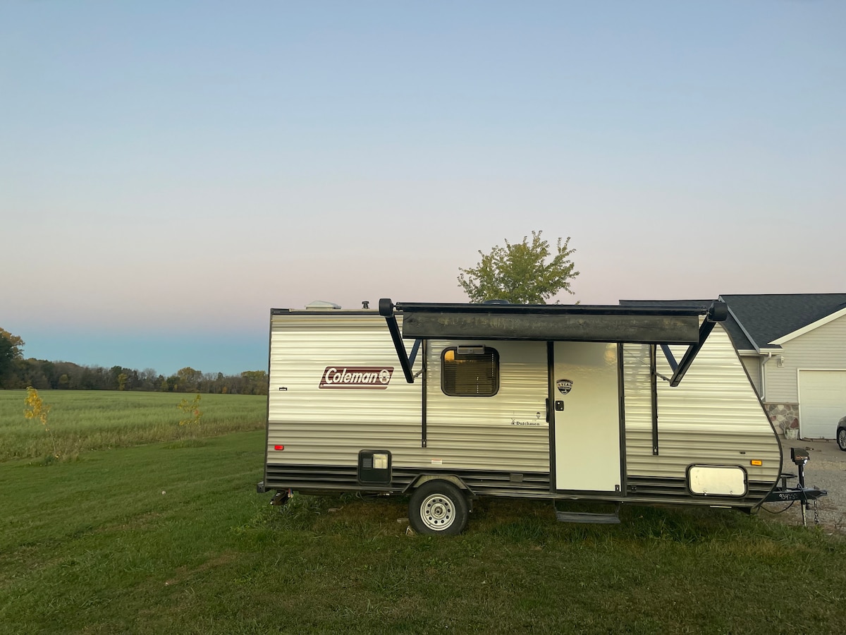 A compact travel trailer is parked on a grassy area, surrounded by expansive fields. The structure features a retractable awning and large windows, providing a view of the serene outdoor landscape. A peaceful evening sky transitions from light to darker hues in the background.