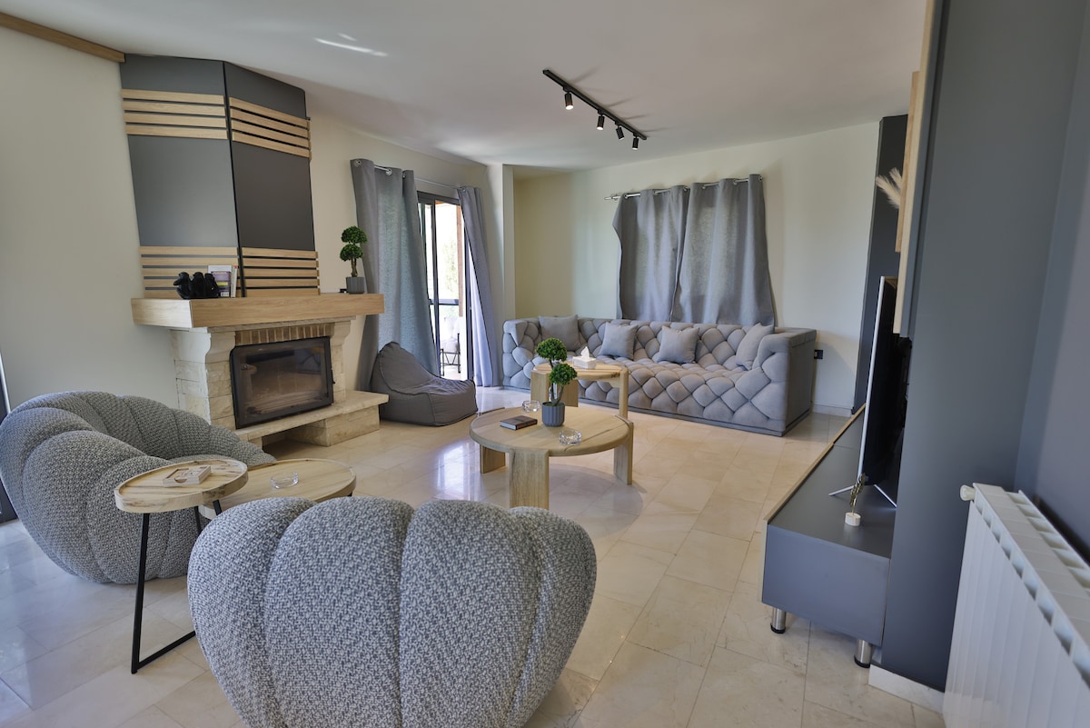 A spacious living area is presented, featuring a modern design with light-colored walls and stone flooring. A comfortable sofa and distinctive armchairs are arranged around a wooden coffee table. Natural light filters through large windows, highlighting the fireplace and decorative plants.