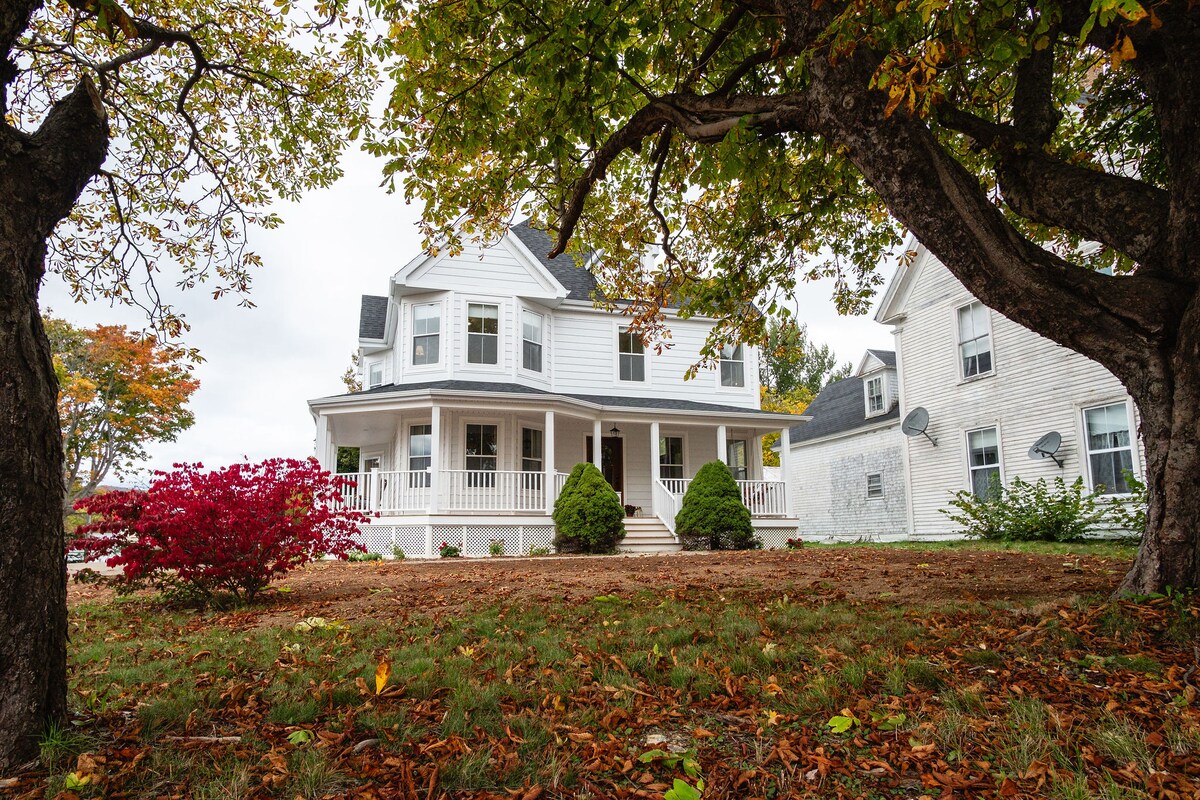 A charming Victorian home is framed by trees with colorful autumn leaves. The exterior features a wide porch with white railing, bordered by manicured shrubs. Vibrant red foliage adds a pop of color to the well-kept lawn, creating an inviting outdoor space.