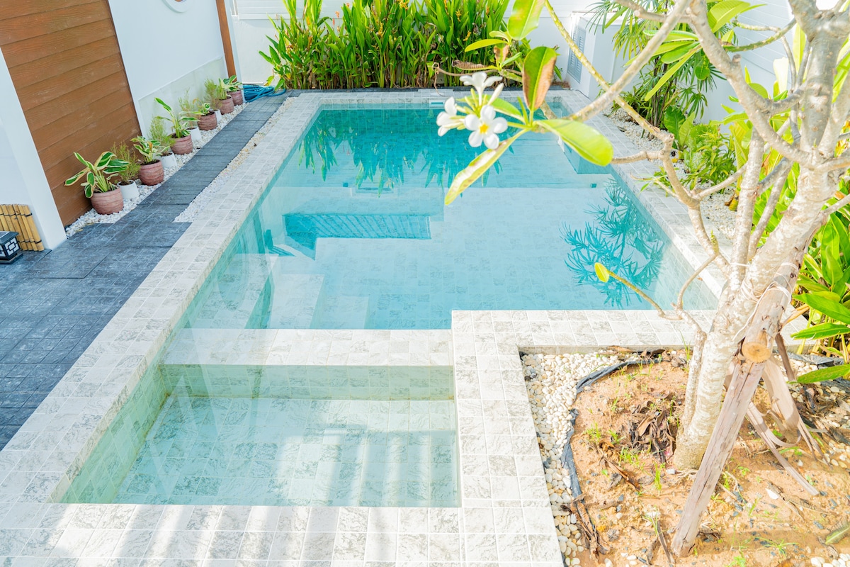 A private swimming pool is surrounded by well-maintained greenery and tropical plants. The pool features a shallow section and is bordered by light-colored stone tiles. Sunlight reflects off the water's surface, creating a serene environment.
