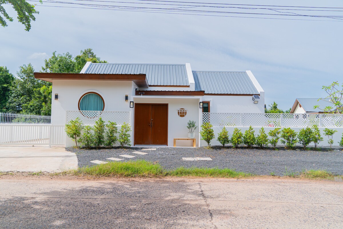 The exterior of the villa is shown, featuring a blend of white walls and a modern roof. A round window adds a unique architectural element. A well-maintained garden with small bushes and a gravel walkway leads to the entrance, offering a sense of privacy.