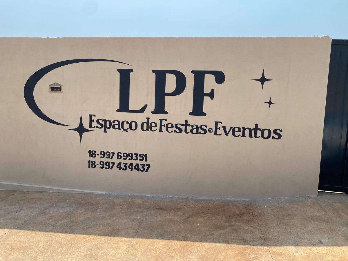 A large, painted wall displays the name 'LPF Espaço de Festas e Eventos' in bold black lettering. Contact numbers are included below the name. The background is a neutral color, with a clear, unobstructed sky visible above.