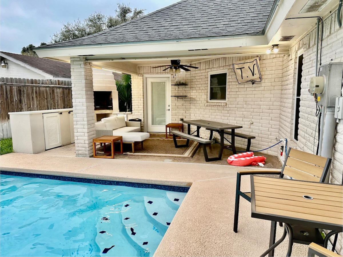 A private outdoor area features a swimming pool and a covered seating space. A wooden dining table is accompanied by several chairs, while a lounge area with sofa seating is present. A ceiling fan is mounted above, and a TV is affixed to the wall, providing entertainment options.